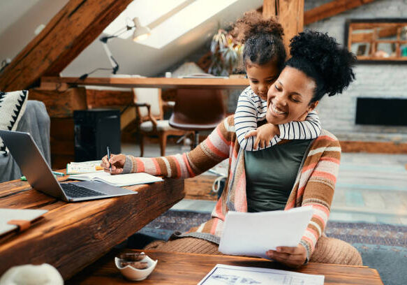 Happy African American mother using laptop and working on paperwork while her daughter is embracing her at home.